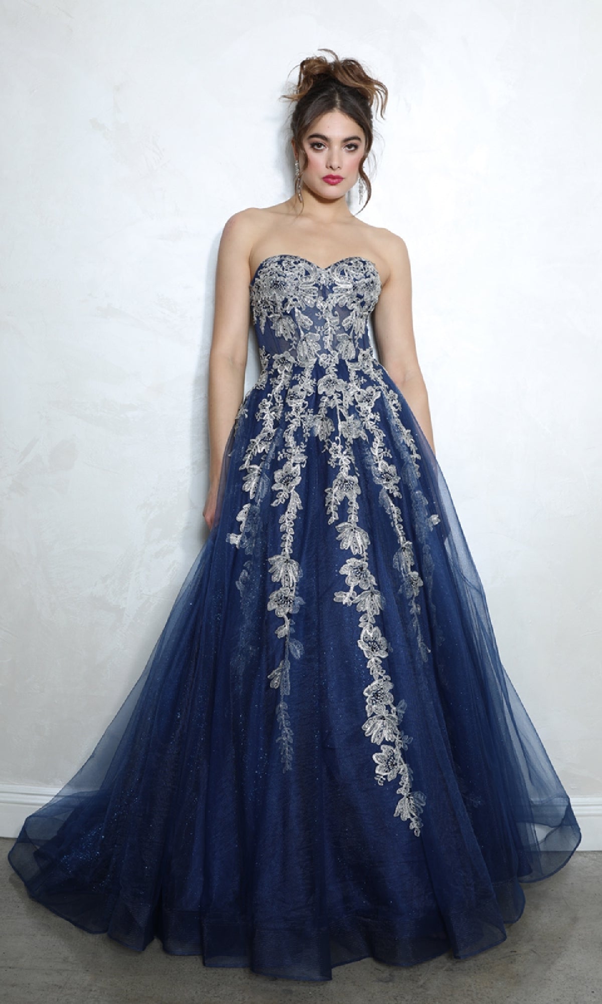 A woman stands elegantly against a light-colored wall wearing the Eureka EK115 Long Blue Sweetheart Prom Gown, a navy strapless A-line design with silver floral embroidery.